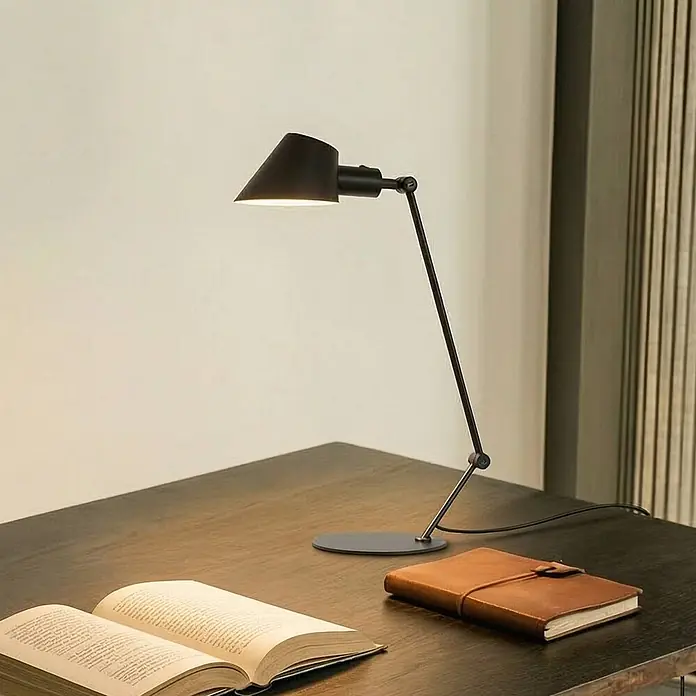 Lindby Tischleuchte Corali - schwarz, schwarz, E27 - Schwarze verstellbare Schreibtischlampe beleuchtet ein offenes Buch auf einem dunklen Holztisch neben einem Ledernotizbuch.