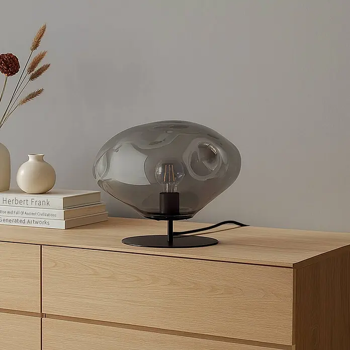 Lindby Tischleuchte Lelia - L x B x H: 35 x 35 x 27,5 cm, alu / grau / zink, E27 - Tischlampe mit rauchgrauem, organischem Glasschirm und schwarzem Fuß auf einer Holzkommode.