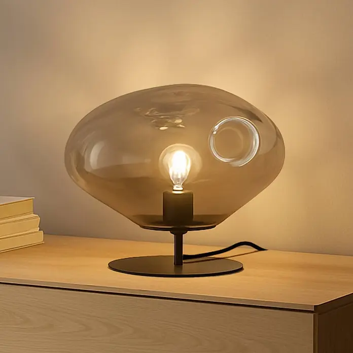 Lindby Tischleuchte Lelia - L x B x H: 35 x 35 x 27,5 cm, alu / grau / zink, E27 - Bernsteinfarbene Glastischlampe mit schwarzem Sockel und sichtbarer Glühbirne, leuchtend auf einer Holzkommode.