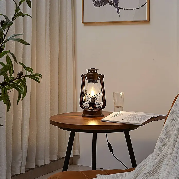 Lindby Tischleuchte Raisa - braun / rost, E14 - Eine beleuchtete Laternenlampe im Vintage-Stil aus dunklem Metall und Glas auf einem Beistelltisch.