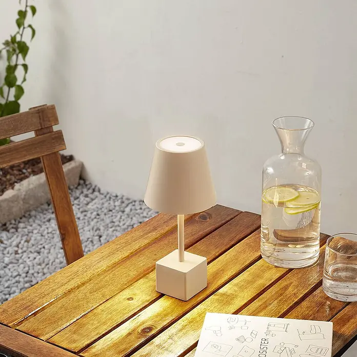 Lindby Tischleuchte Janea - creme / amber, creme / amber - Cremefarbene kabellose Tischlampe auf einem Holztisch im Freien neben einer Karaffe mit Zitronenwasser, die ihre Verwendung im Freien zeigt.