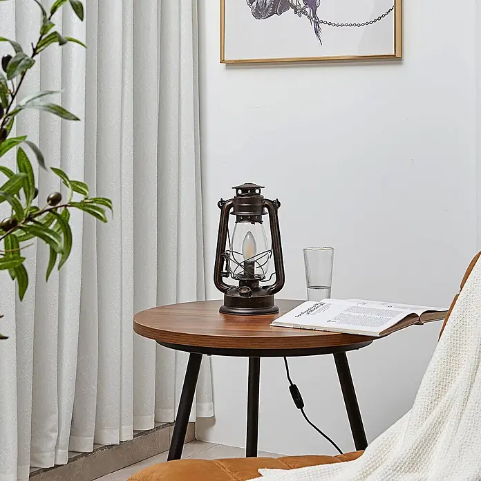 Lindby Tischleuchte Raisa - braun / rost, E14 - Laternen-Tischleuchte in Bronzeoptik beleuchtet einen Beistelltisch, neben einem Stuhl und Buch.
