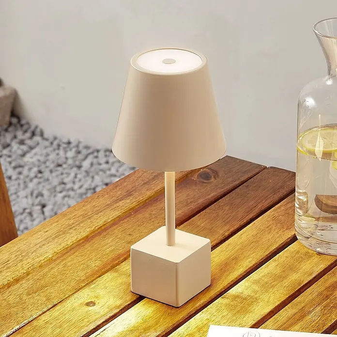 Lindby Tischleuchte Janea - creme / amber, creme / amber - Eine beige, matte Akku-Tischlampe mit konischem Schirm und quadratischem Fuß steht auf einem hölzernen Latten-Außentisch neben einer Glaskaraffe.