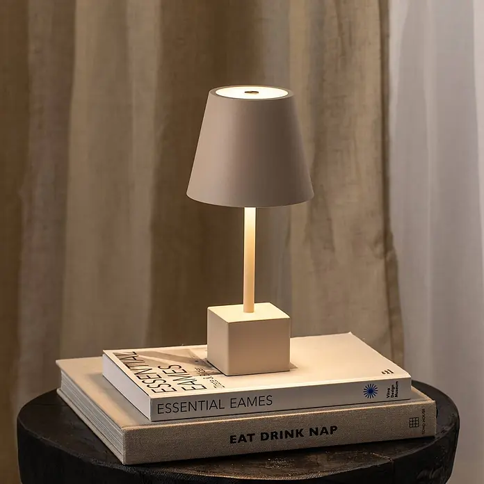 Lindby Tischleuchte Janea - creme / amber, creme / amber - Eine moderne graue Tischlampe mit konischem Schirm, dünnem Stiel und quadratischem Fuß beleuchtet zwei Bücher auf einem dunklen Holztisch.