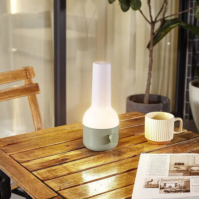 Lindby Tischleuchte Isaline - grün, weiß / opal - Moderne, tragbare Lampe mit weißem Oberteil und salbeigrüner Basis auf einem Holztisch, neben Tasse und Zeitschrift. Ideal für draußen.