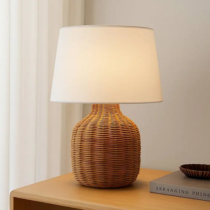 Lindby Tischleuchte Lirenda - L x B x H: 35 x 35 x 52 cm, Holz hell, weiß / opal, E27 - Beleuchtete Tischlampe mit geflochtenem Rattansockel und weißem Stoffschirm auf einem Holztisch.