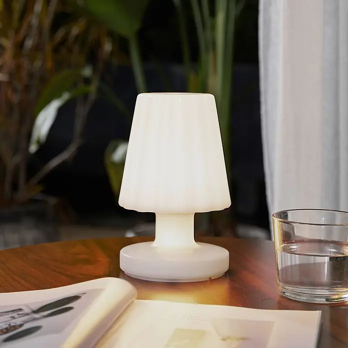 Lindby Tischleuchte Jovem - weiß / opal, weiß / opal - Weiße beleuchtete glockenförmige Tischlampe auf einem Holztisch neben einem Buch und Glas.