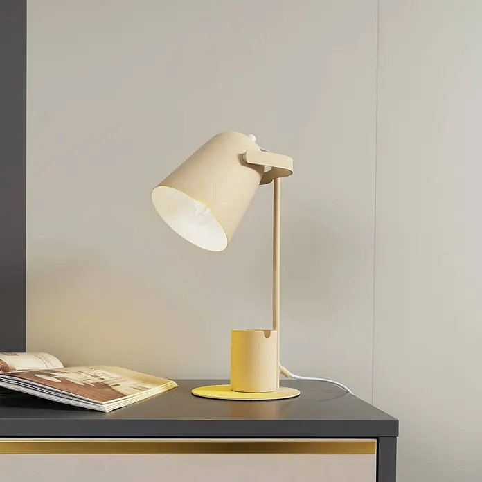 Lindby Tischleuchte Ilmar - L x B x H: 20 x 15 x 38 cm, creme / amber, creme / amber, E27 - Beige Metall-Schreibtischlampe mit integriertem Stifthalter, beleuchtet ein offenes Magazin auf einem dunklen Schreibtisch.