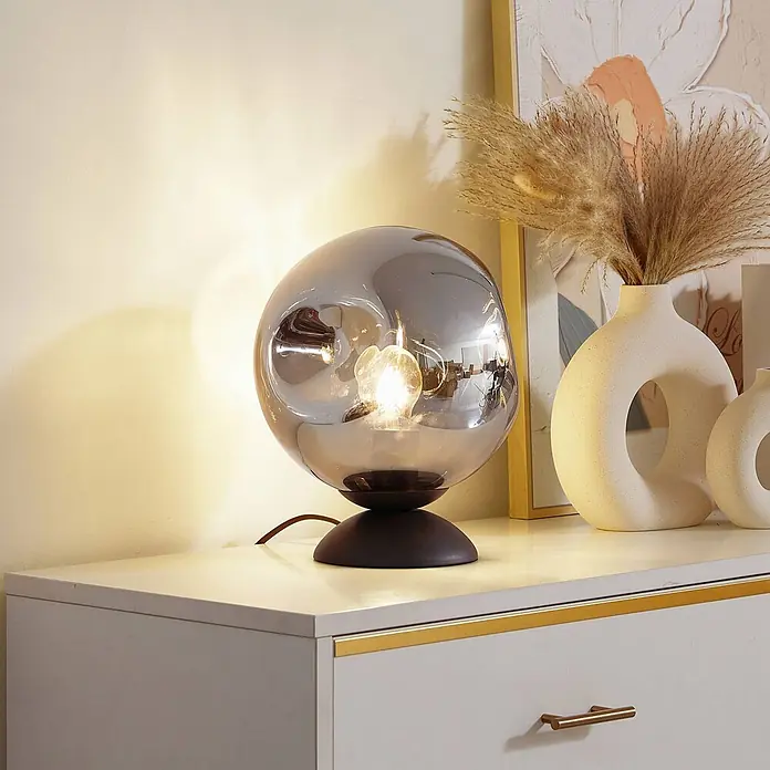 Lindby Tischleuchte Valentina - L x B x H: 25 x 25 x 30 cm, braun / rost, alu / grau / zink, E27 - Beleuchtete Tischlampe mit rauchgrauem Glasschirm und dunklem Sockel auf einer weißen Kommode neben Ziergefäßen.