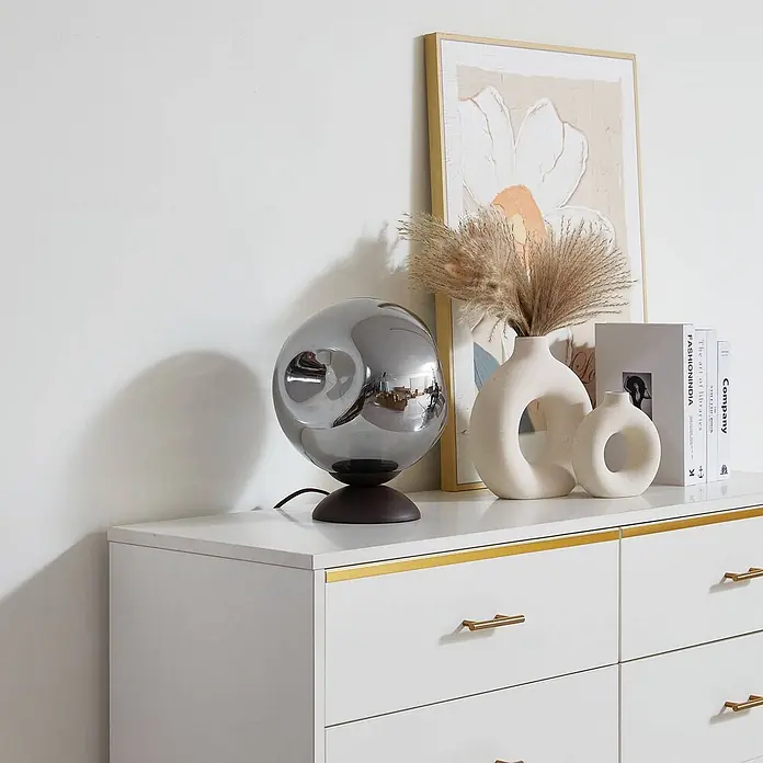 Lindby Tischleuchte Valentina - L x B x H: 25 x 25 x 30 cm, braun / rost, alu / grau / zink, E27 - Rauchgraue kugelförmige Glastischlampe mit dunklem Sockel auf weißer Kommode.