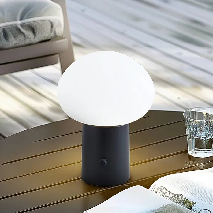 Lindby Tischleuchte Zelie - schwarz, weiß / opal - Schwarze und weiße tragbare Tischlampe mit kugelförmigem Diffusor, die ein warmes Licht auf einen Holztisch im Freien neben einem Stuhl wirft.