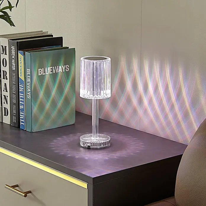 Lindby Tischleuchte Louane - L x B x H: 8,5 x 8,5 x 25,7 cm, klar / transparent, klar / transparent - Transparente Acryl-Tischlampe mit Rillen, die bunte Lichtmuster auf einen Tisch neben Büchern wirft.