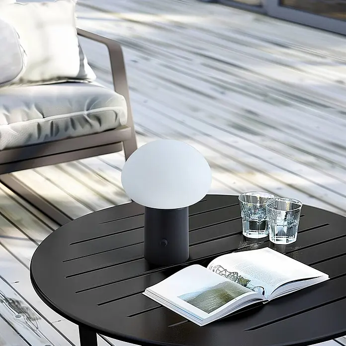 Lindby Tischleuchte Zelie - schwarz, weiß / opal - Tragbare Lampe mit weißem kugelförmigem Schirm und dunklem zylindrischem Fuß auf einem Outdoor-Lamellentisch mit Buch und Gläsern.