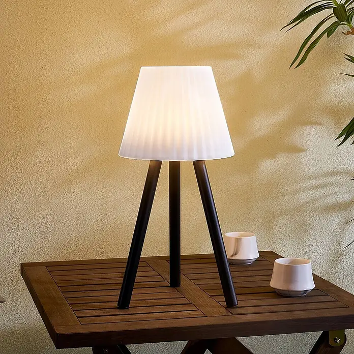 Lindby Tischleuchte Jaimy - schwarz, weiß / opal - Weiße plissierte Tischlampe mit dunklen Holz-Dreibeinbeinen, auf einem Outdoor-Lamellentisch mit Keramiktassen.