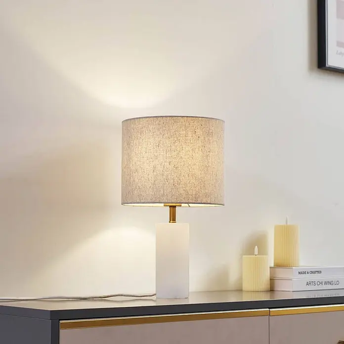 Lindby Tischleuchte Alabaster - L x B x H: 25 x 25 x 43 cm, weiß / opal, creme / amber, E27 - Eine Tischleuchte mit grauem Leinenschirm und weißem Sockel beleuchtet eine Kommode neben Büchern und Kerzen.