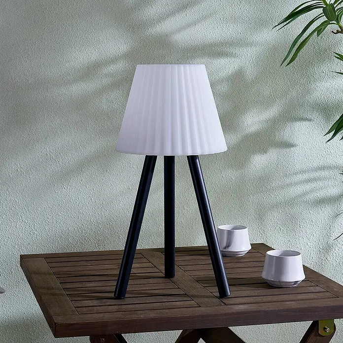 Lindby Tischleuchte Jaimy - schwarz, weiß / opal - Eine Tischlampe mit weißem Plisseeschirm und schwarzen Dreibeinbeinen steht auf einem Holzlamellentisch neben zwei weißen Tassen.