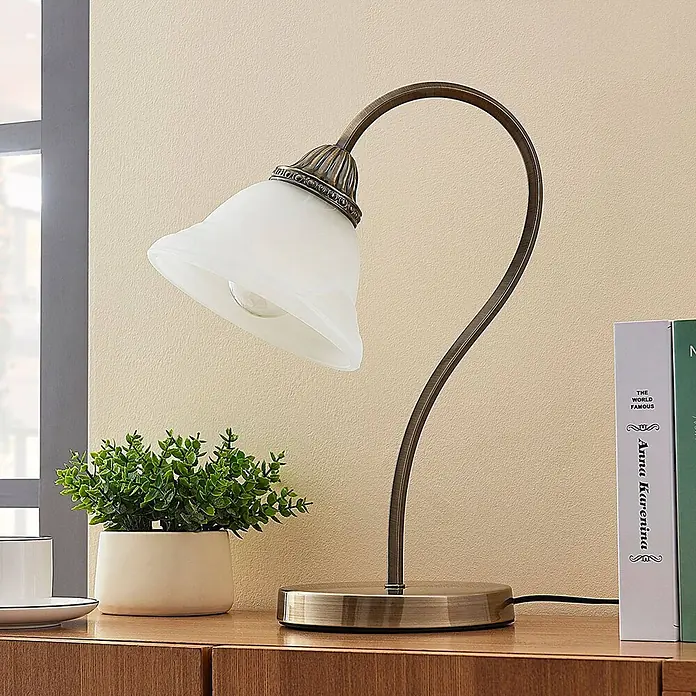 Lindby Tischleuchte Mialina - bronze / altmessing, E27 - Eine antik messingfarbene Tischlampe mit weißem, geriffeltem Glasschirm steht auf einem Holzschreibtisch neben einer Topfpflanze.