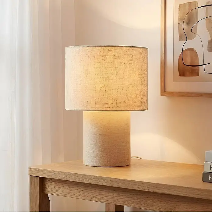 Lindby Tischleuchte Elfie - creme / amber, E27 - Moderne Tischlampe mit zylindrischem, texturiertem hellbeigem Sockel und passendem Stoffschirm, die einen hellen Holztisch beleuchtet.