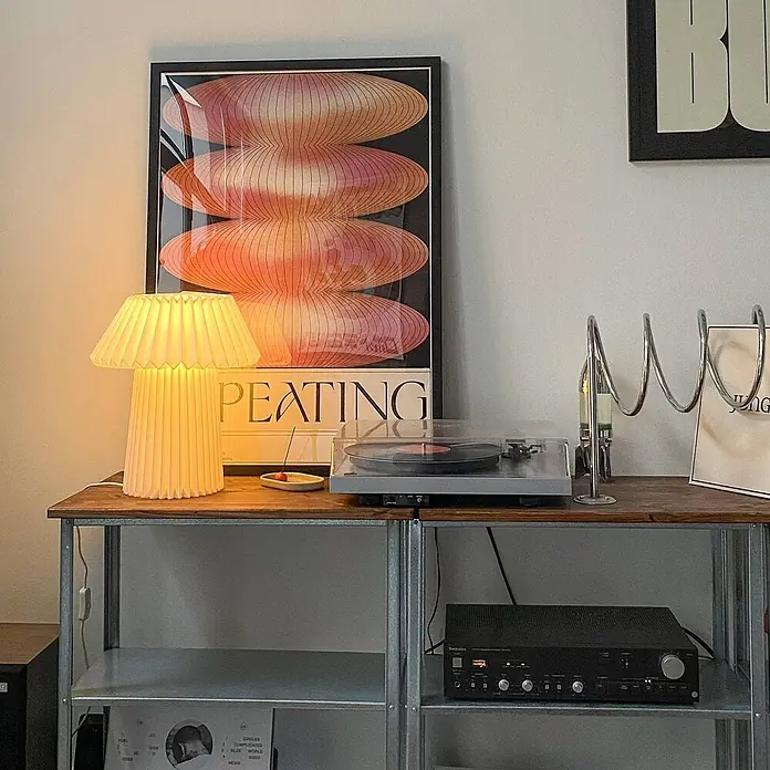 Lindby Tischleuchte Magali - weiß / opal, E14 - Stilvolles Audio-Setup: Plattenspieler, schwarzer Verstärker und eine warme plissierte Lampe auf einem Holzregal.