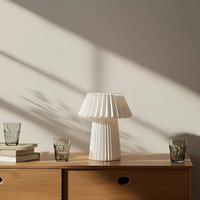 Lindby Tischleuchte Magali - weiß / opal, E14 - Weiße plissierte Tischlampe auf einer Holzkommode, umgeben von Gläsern und Büchern.