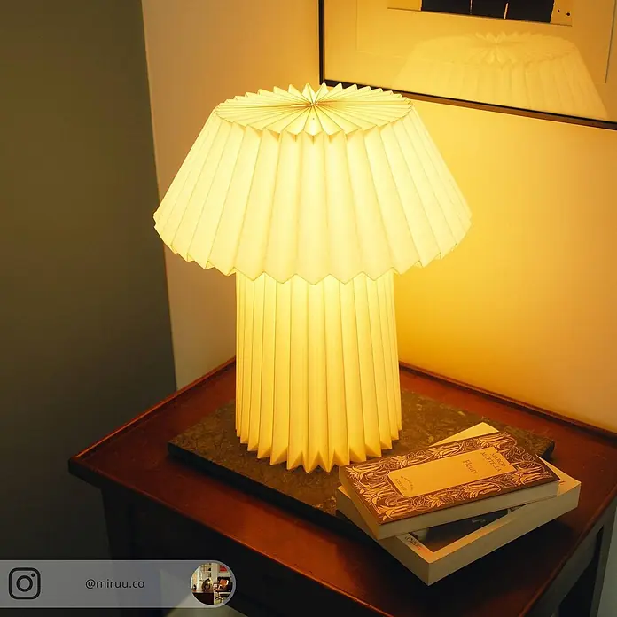 Lindby Tischleuchte Magali - weiß / opal, E14 - Eine warm leuchtende plissierte Papier-Tischlampe erhellt einen hölzernen Nachttisch mit gestapelten Büchern.