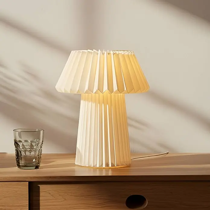 Lindby Tischleuchte Magali - weiß / opal, E14 - Eine cremefarbene, plissierte Tischlampe mit warmem Licht auf einer Holzkommode neben einem Glas.