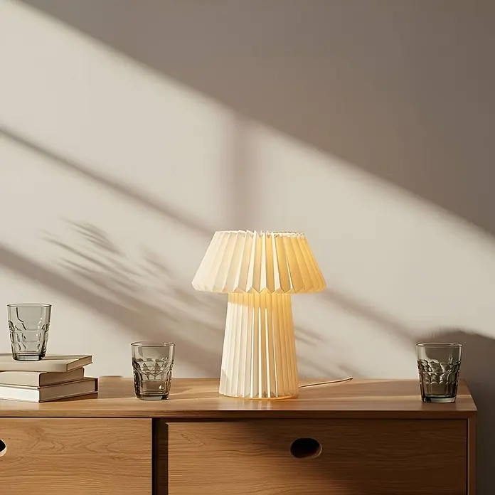 Lindby Tischleuchte Magali - weiß / opal, E14 - Eine beleuchtete cremefarbene Plissee-Tischlampe steht auf einer Holzkommode, umgeben von Gläsern und Büchern.