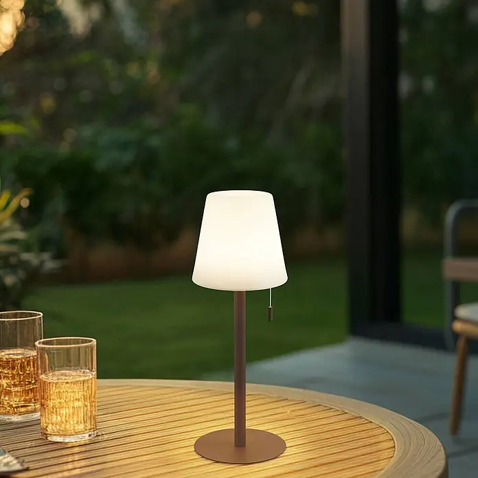 Lindby Tischleuchte Azalea - L x B x H: 16,5 x 16,5 x 45 cm, schwarz - Bronze Tischlampe mit weißem konischem Schirm auf einem hölzernen Außentisch, neben zwei Gläsern mit Getränken.