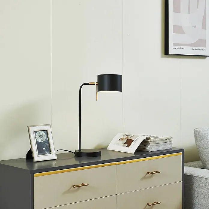 Lindby Tischleuchte Kaida - L x B x H: 30 x 15 x 43,5 cm, schwarz, schwarz, E27 - Schwarze Tischleuchte mit Goldakzenten auf einer Kommode, beleuchtet, neben Fotorahmen und Zeitschriften.