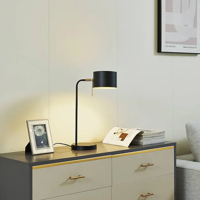 Lindby Tischleuchte Kaida - L x B x H: 30 x 15 x 43,5 cm, schwarz, schwarz, E27 - Elegante schwarze Tischlampe mit goldenen Akzenten, die warmes Licht auf eine Kommode wirft.