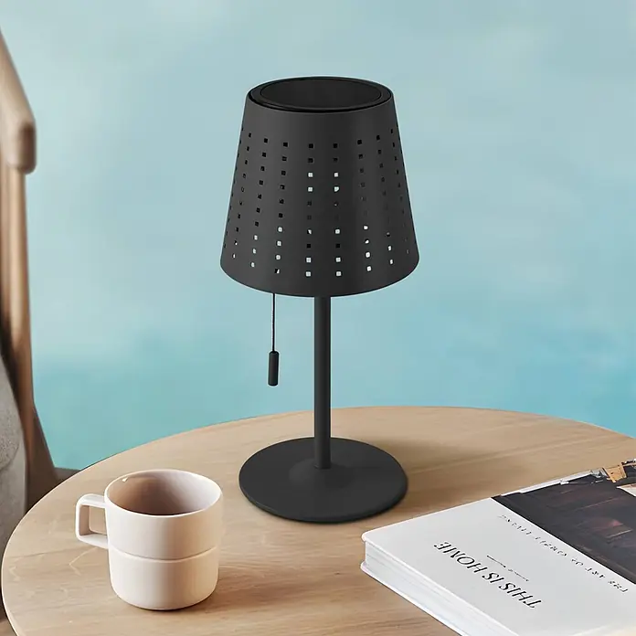 Lindby Tischleuchte Hilario - schwarz, schwarz - Schwarze perforierte Tischlampe mit Zugschalter auf hellem Holztisch neben einer Tasse und einem Buch.