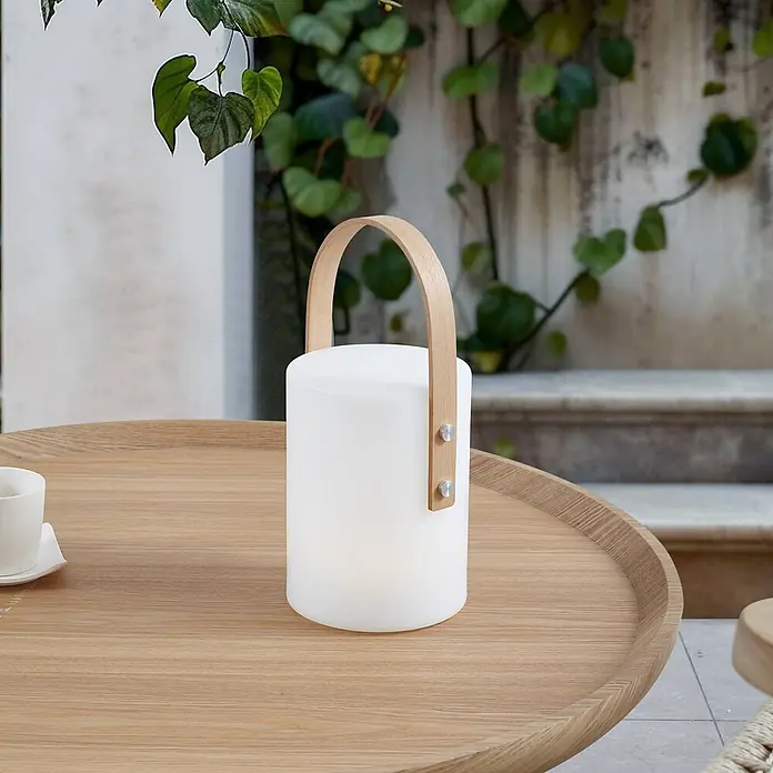 Lindby Tischleuchte Juria - L x B x H: 16,5 x 16,5 x 35,5 cm, weiß / opal - Beleuchtete weiße zylindrische Laterne mit hellem Holzgriff auf einem Holztisch im Freien.