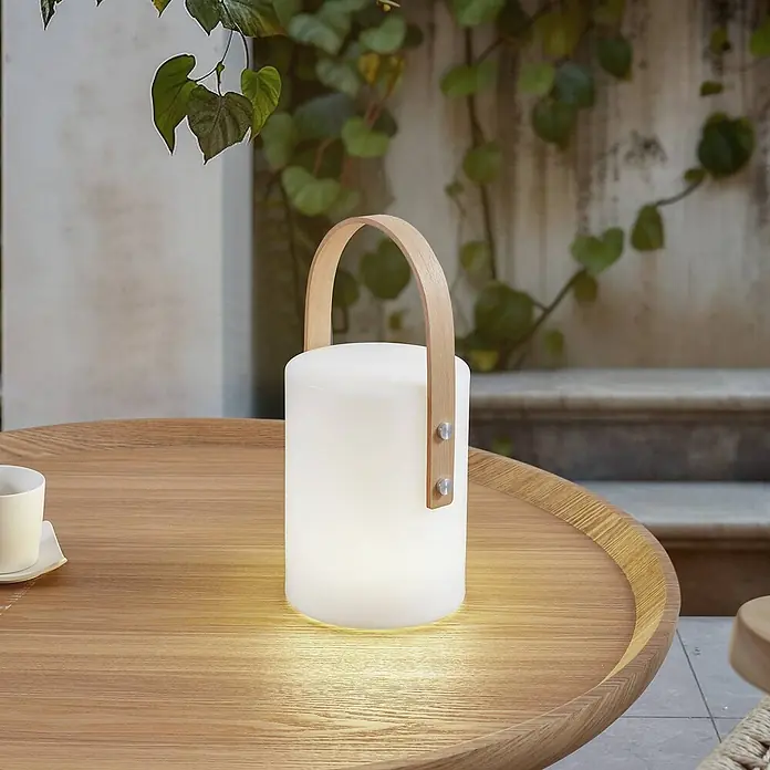 Lindby Tischleuchte Juria - L x B x H: 16,5 x 16,5 x 35,5 cm, weiß / opal - Eine leuchtende weiße Zylinderlampe mit hellem Holzgriff steht auf einem hölzernen Gartentisch. Grüne Ranken sind im Hintergrund.