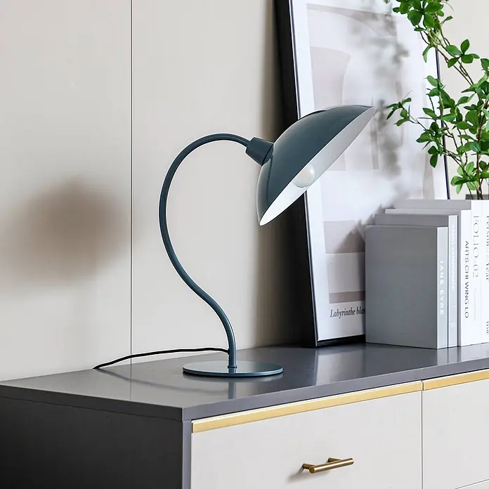 Lucande Tischleuchte Arvadon - blau, blau, E14 - Türkisfarbene Schreibtischlampe mit gebogenem Arm und weitem Schirm, auf einem dunklen Schrank neben Büchern.
