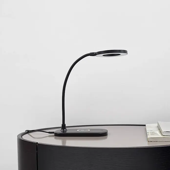 Lindby Tischleuchte Siona - schwarz, schwarz - Schwarze flexible LED-Tischlampe mit rundem Kopf und kabelloser Ladefunktion auf dunklem Beistelltisch neben Büchern.