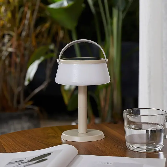 Lindby Tischleuchte Mikkel - L x B x H: 14,2 x 14,2 x 7,2 cm, creme / amber - Tragbare hellbeige Tischlampe mit Griff, auf einem Holztisch neben einem Wasserglas und einem Buch.