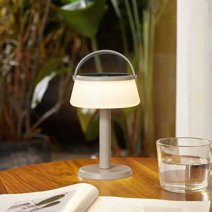 Lindby Tischleuchte Mikkel - L x B x H: 14,2 x 14,2 x 7,2 cm, creme / amber - Eine leuchtende, hellgraue tragbare Lampe mit einem durchscheinenden weißen Schirm und Solarpanel steht auf einem Holztisch neben einem Buch und Wasserglas.
