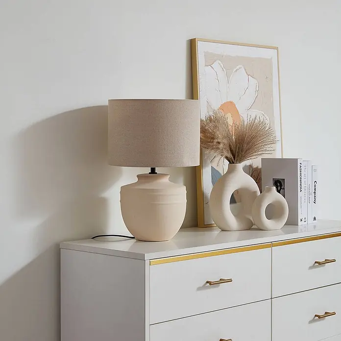 Lindby Tischleuchte Thalassia - L x B x H: 30 x 30 x 45,5 cm, creme / amber, creme / amber, E27 - Eine beige Keramiktischlampe mit hellbraunem Stoffschirm steht auf einer weißen Kommode mit goldenen Akzenten.