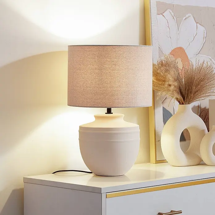 Lindby Tischleuchte Thalassia - L x B x H: 30 x 30 x 45,5 cm, creme / amber, creme / amber, E27 - Eine cremefarbene Keramik-Tischlampe mit hellbraunem Stoffschirm auf einer weißen Kommode. Die Lampe beleuchtet den Bereich warm.