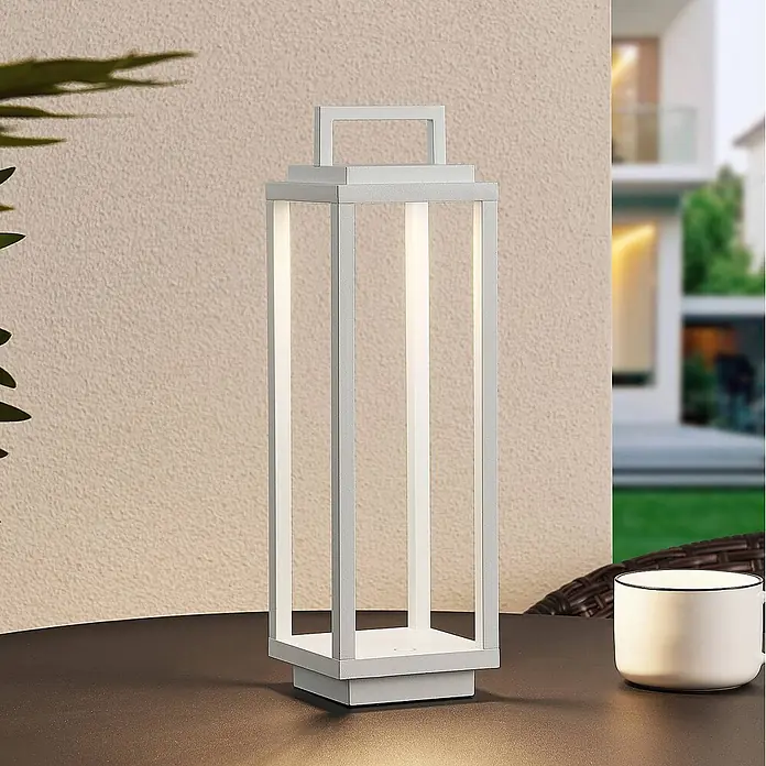 Lucande Tischleuchte Mirina - L x B x H: 10 x 10 x 33,5 cm, weiß / opal - Weiße LED-Laternen-Tischleuchte mit Griff, beleuchtet, auf einem dunklen Outdoor-Tisch neben einer weißen Tasse.