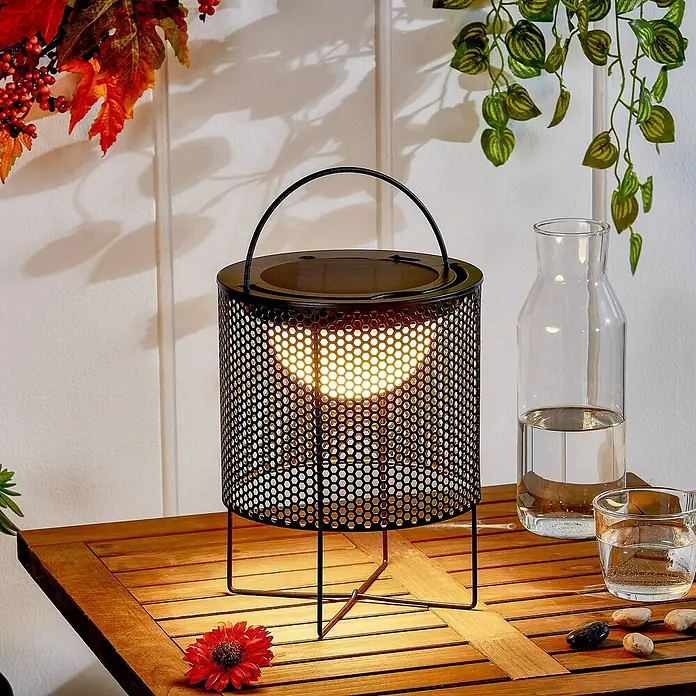 Lindby Tischleuchte Achilleas - L x B x H: 20 x 20 x 26 cm, schwarz - Schwarze Metall-Laternen-Tischlampe mit sechseckiger Perforation und beleuchtetem Inneren, auf einem Holztisch.