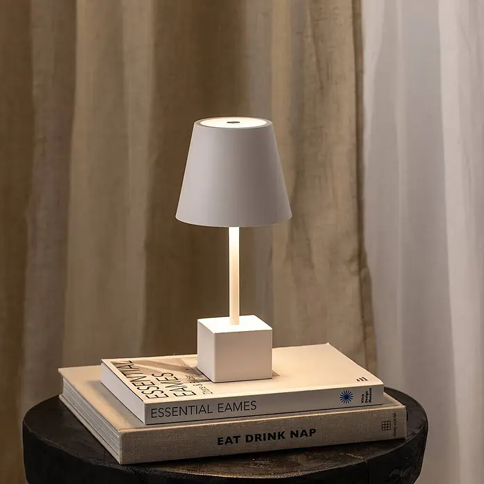Lindby Tischleuchte Janea - weiß / opal, weiß / opal - Moderne graue Tischlampe mit weißem Sockel, die warmes Licht abstrahlt. Sie steht auf gestapelten Büchern auf einem dunklen Holzhocker.