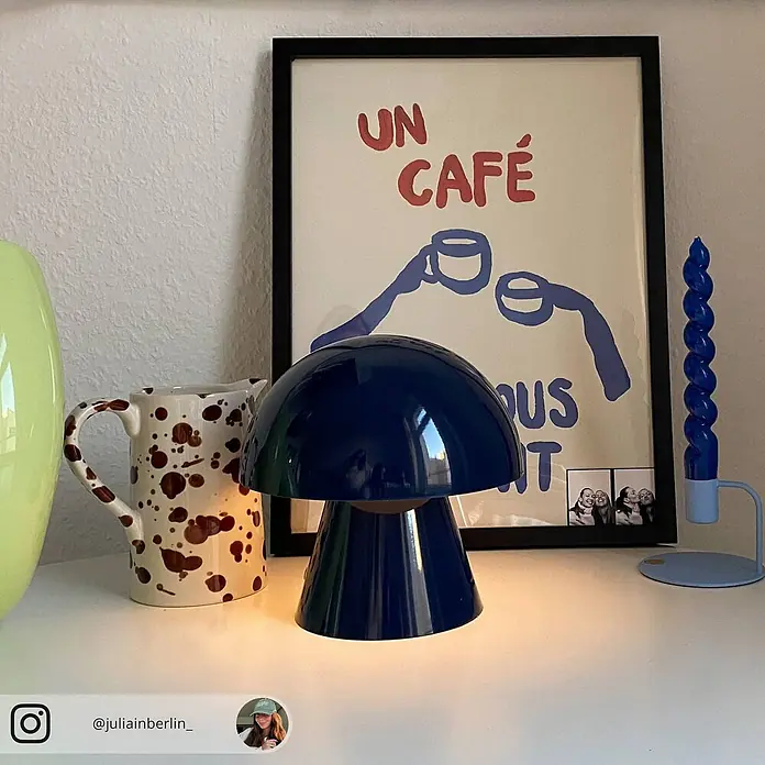 Lindby Tischleuchte Nevijo - L x B x H: 18 x 18 x 17,5 cm, blau, blau - Eine glänzende, dunkelblaue Pilz-Tischlampe beleuchtet eine Oberfläche auf einem dekorierten Regal, neben einem gesprenkelten Krug und einem gerahmten Poster.