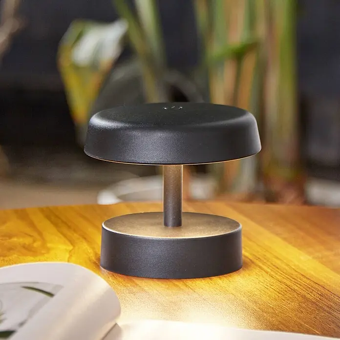 Lindby Tischleuchte Nalani - schwarz, schwarz - Minimalistische schwarze und bronzefarbene tragbare Tischlampe spendet warmes Licht auf einer Holzoberfläche.
