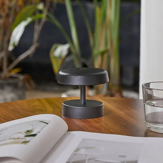 Lindby Tischleuchte Nalani - schwarz, schwarz - Schwarze, minimalistische Tischlampe auf einem Holztisch neben einem geöffneten Buch und einem Glas Wasser.