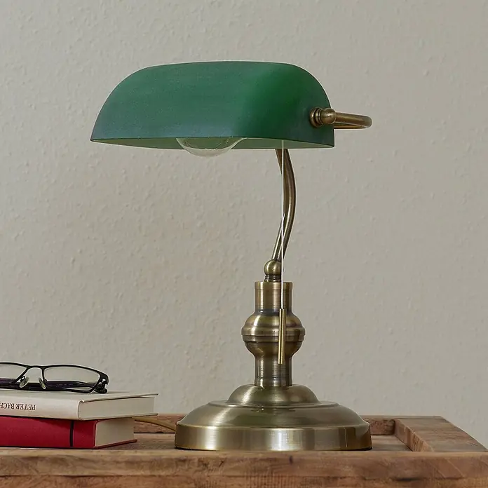 Lindby Tischleuchte Milenka - L x B x H: 18,5 x 23 x 38 cm, bronze / altmessing, grün, E27 - Messing-Bankerlampe mit grünem Schirm auf einem Holztisch neben Büchern und Brille.