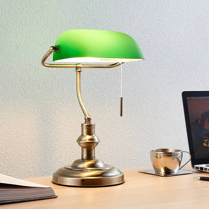 Lindby Tischleuchte Milenka - L x B x H: 18,5 x 23 x 38 cm, bronze / altmessing, grün, E27 - Klassische Bankerlampe mit grünem Schirm und antikem Messingfuß, beleuchtet einen Holzschreibtisch neben Laptop und Buch.