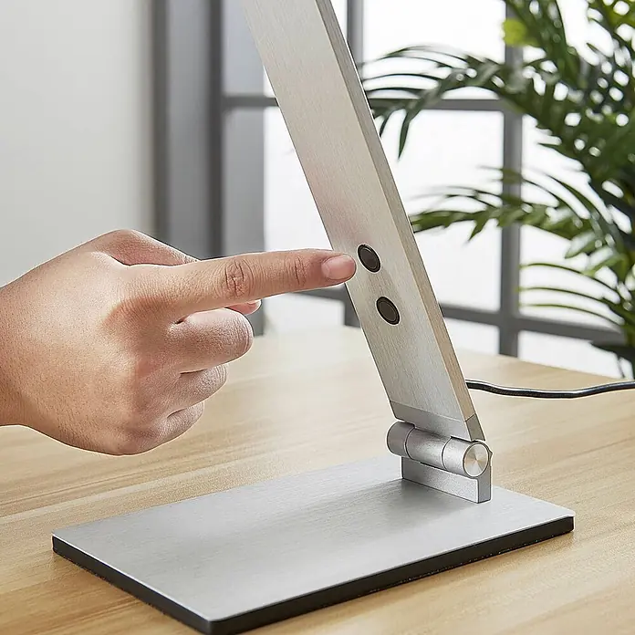 Lucande Tischleuchte Nicano - L x B x H: 34,5 x 12 x 39 cm, alu / grau / zink, alu / grau / zink - Eine Hand drückt einen Knopf auf einer silbernen, gebürsteten Aluminium-Schreibtischlampe auf einem Holztisch.