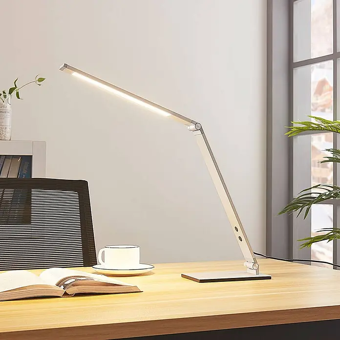 Lucande Tischleuchte Nicano - L x B x H: 34,5 x 12 x 39 cm, alu / grau / zink, alu / grau / zink - Eine moderne, verstellbare silberne LED-Schreibtischlampe beleuchtet einen Holztisch mit Buch und Tasse.