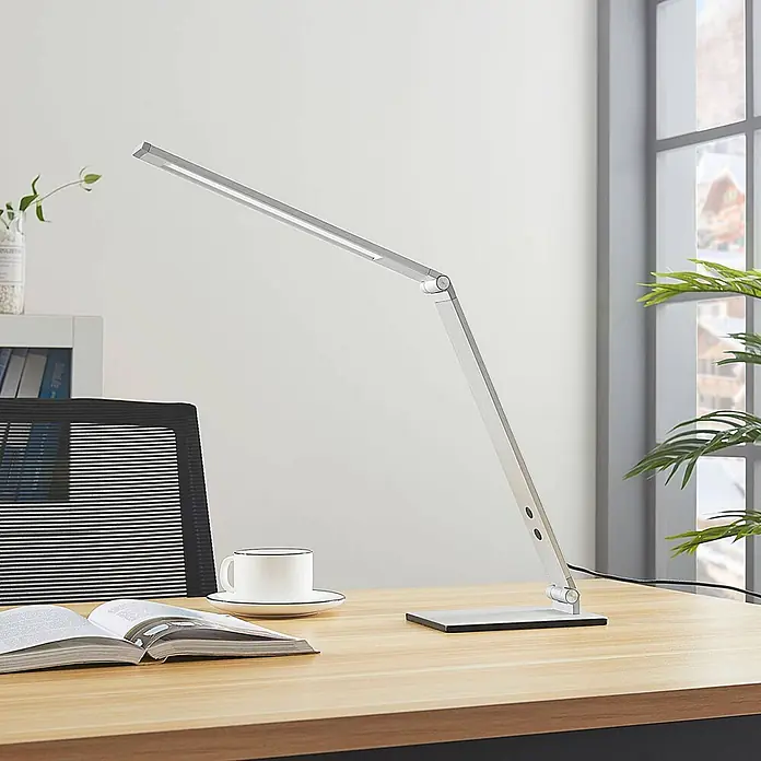 Lucande Tischleuchte Nicano - L x B x H: 34,5 x 12 x 39 cm, alu / grau / zink, alu / grau / zink - Schlanke silberne LED-Schreibtischlampe mit verstellbarem Arm auf einem Holztisch mit offenem Buch und Kaffeetasse.